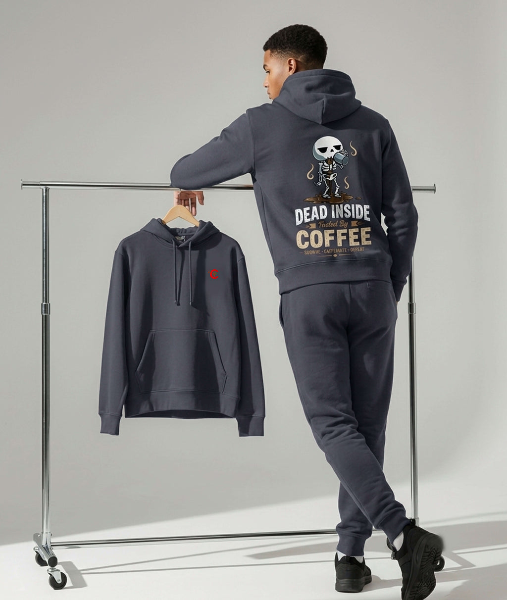 A model wearing a dark gray hoodie and matching sweatpants stands beside a clothing rack in a studio. The hoodie has a large back graphic with a skeleton and the text 'DEAD INSIDE COFFEE', and the matching pants are shown hanging on the rack.