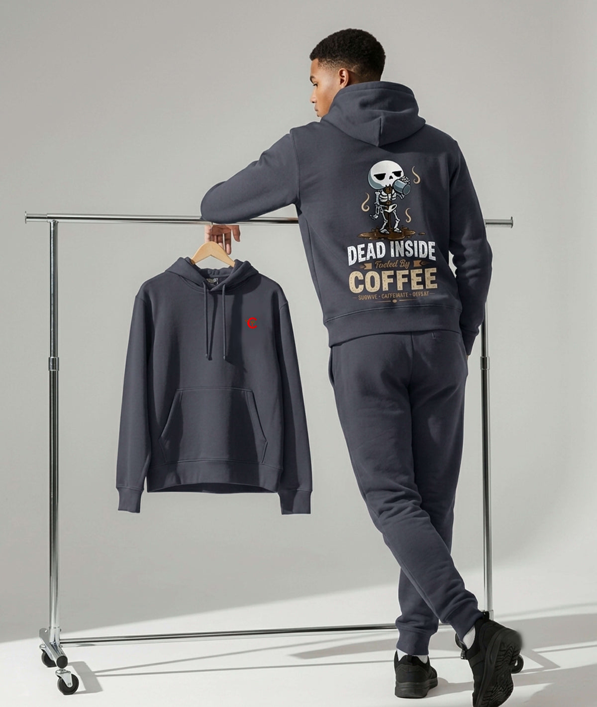 A model wearing a dark gray hoodie and matching sweatpants stands beside a clothing rack in a studio. The hoodie has a large back graphic with a skeleton and the text 'DEAD INSIDE COFFEE', and the matching pants are shown hanging on the rack.