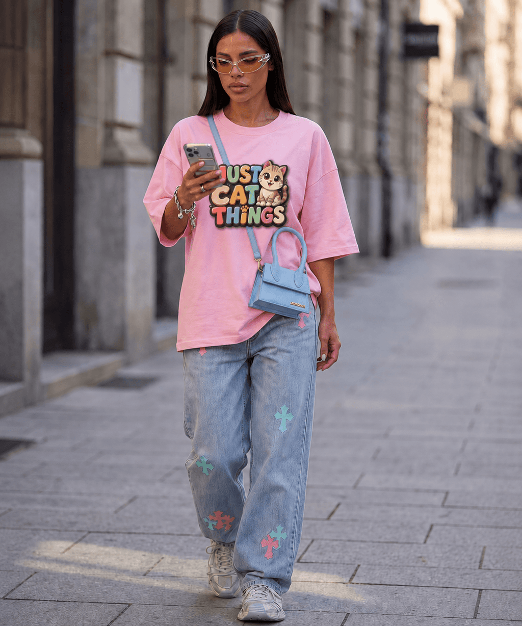 A woman walking down a city street wearing a pink graphic t-shirt with a cartoon bear and the text 'JUST CAT ALL THINGS', light-wash blue jeans with colorful cat prints, and a small light blue crossbody bag.