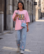 A woman walking down a city street wearing a pink graphic t-shirt with a cartoon bear and the text 'JUST CAT ALL THINGS', light-wash blue jeans with colorful cat prints, and a small light blue crossbody bag.