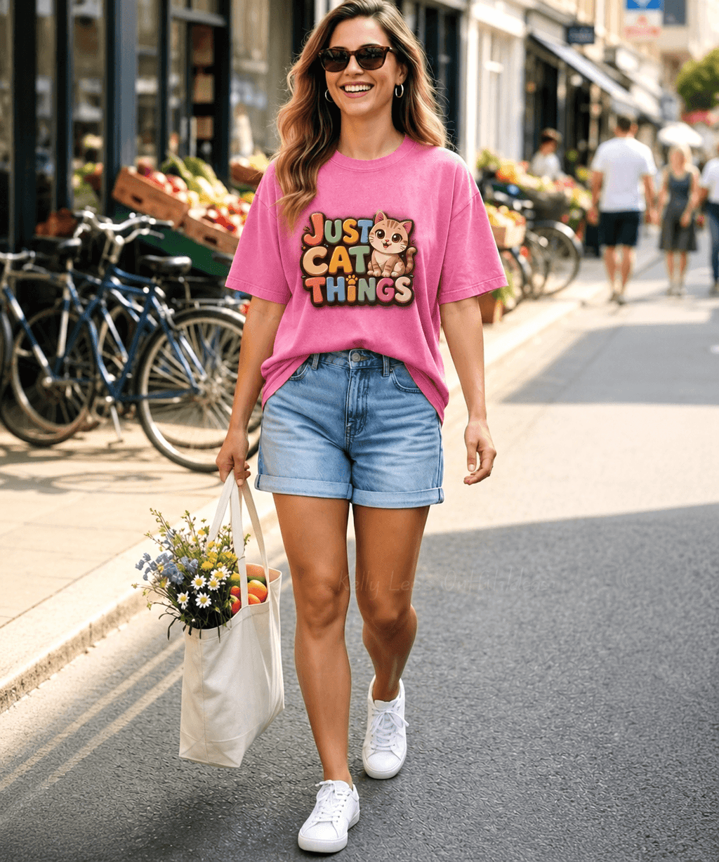 A woman walking on a city street wearing a pink graphic T-shirt that reads 'Just Cat Things' with a cat illustration, light blue denim shorts, white sneakers, and sunglasses, while carrying a tote bag of flowers.