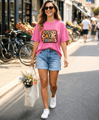 A woman walking on a city street wearing a pink graphic T-shirt that reads 'Just Cat Things' with a cat illustration, light blue denim shorts, white sneakers, and sunglasses, while carrying a tote bag of flowers.
