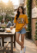 A woman stands at an outdoor cafe table wearing a mustard yellow graphic T-shirt that reads 'JUST CAT THINGS' with a cat illustration, paired with denim shorts and white sneakers.