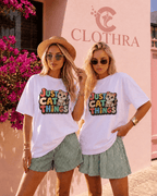 Two women modeling matching white graphic t-shirts with a colorful 'Just Cat Things' print, paired with green and white striped shorts, standing in front of a pink wall with the brand name Clothra.