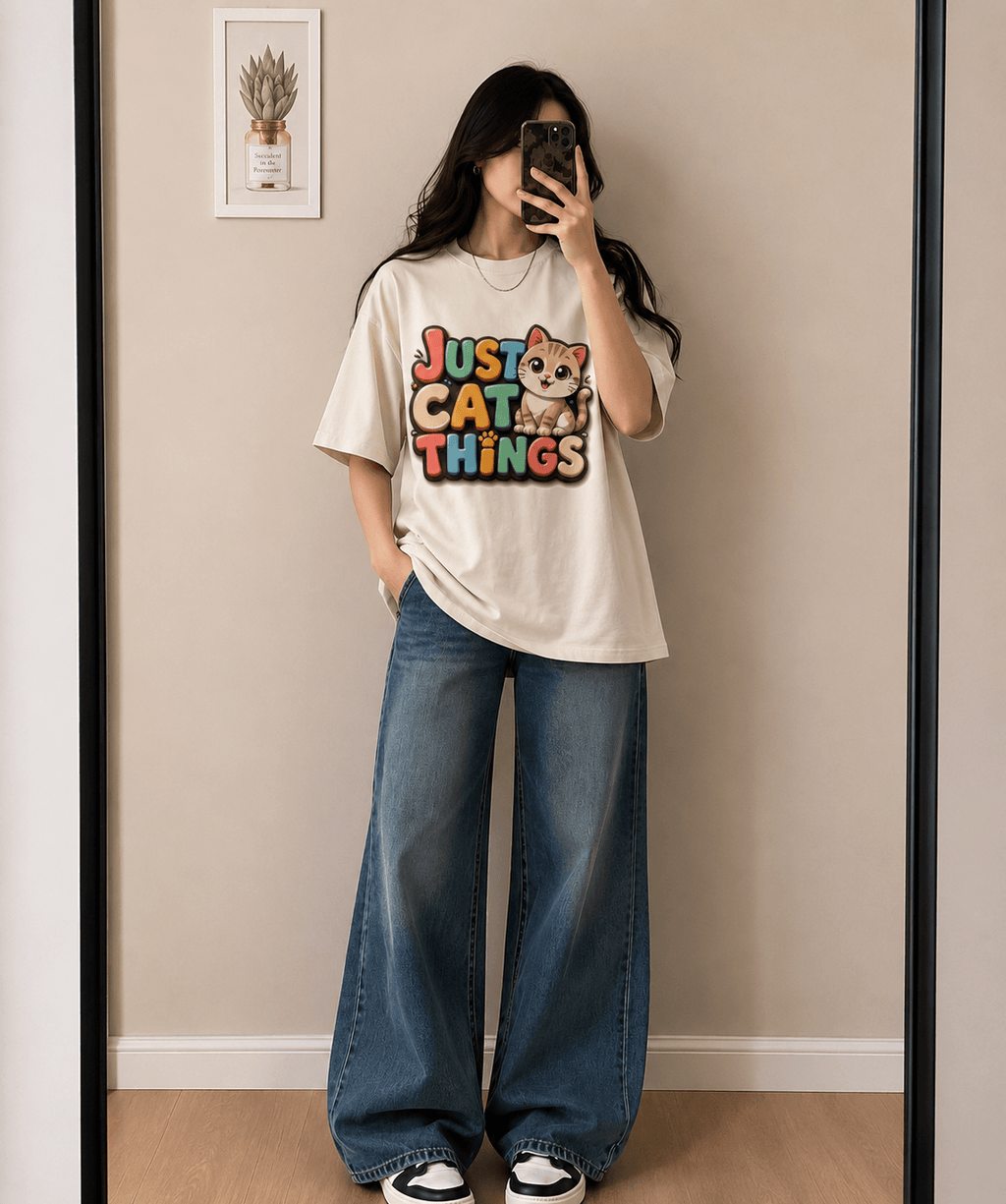 A woman takes a mirror selfie wearing a cream-colored graphic t-shirt with a colorful 'Just Cat Things' print and wide-leg blue jeans.