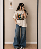 A woman takes a mirror selfie wearing a cream-colored graphic t-shirt with a colorful 'Just Cat Things' print and wide-leg blue jeans.