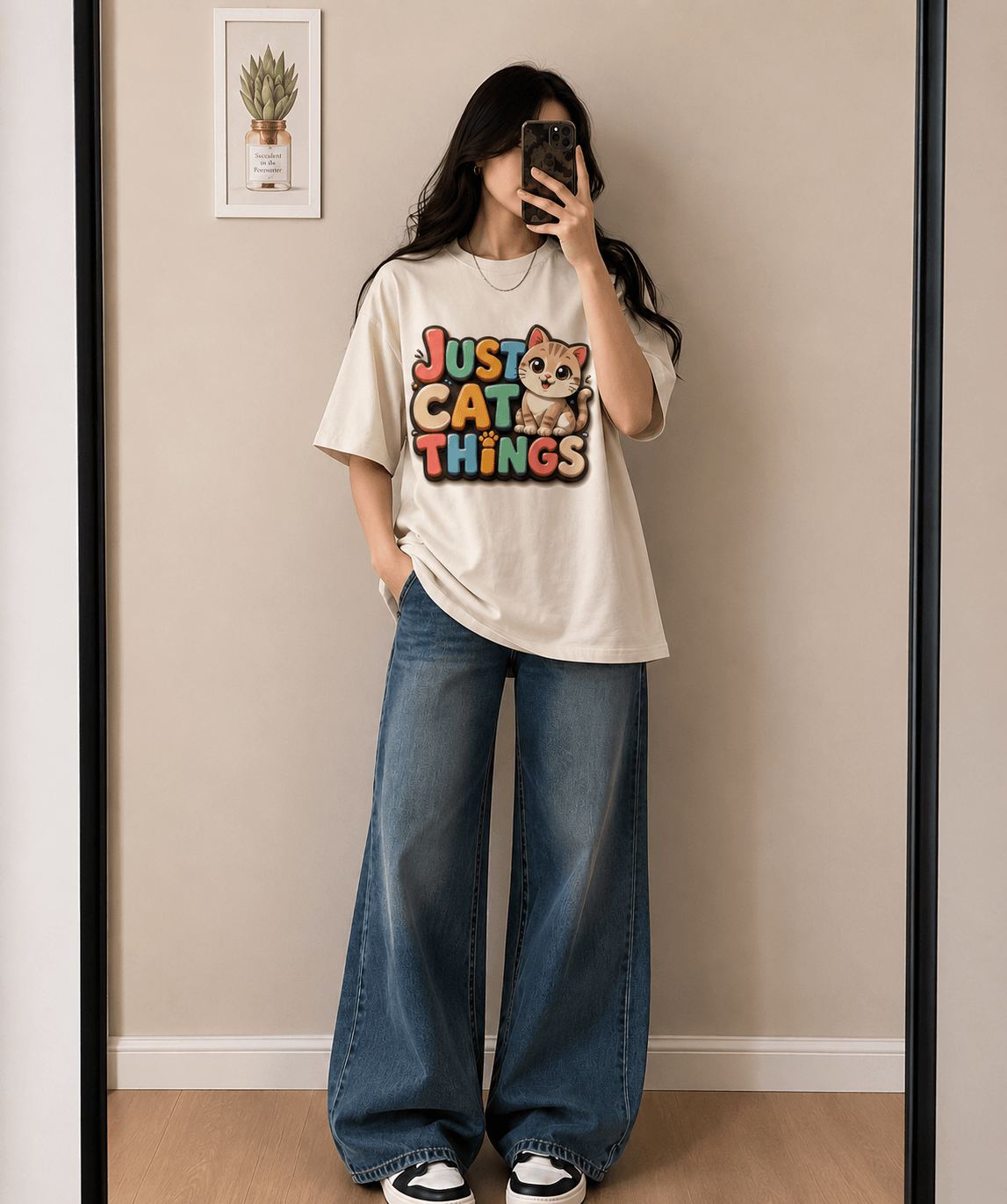 A woman takes a mirror selfie wearing a cream-colored graphic t-shirt with a colorful 'Just Cat Things' print and wide-leg blue jeans.