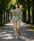 A woman walking down a tree-lined path wearing an oversized sage green graphic t-shirt that reads 'JUST CAT THINGS' with a cat illustration, paired with beige cargo pants and gray sneakers.