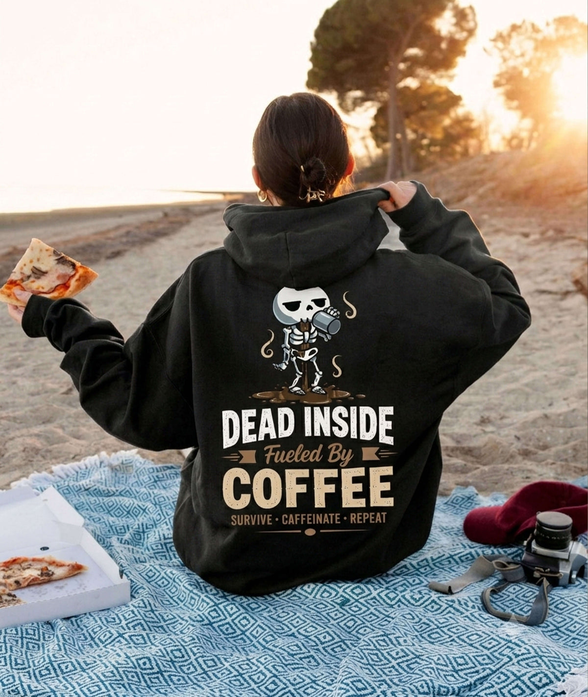 A person sitting on a blue blanket at a beach during sunset, wearing a black hoodie with a skeleton coffee cup graphic and the text 'DEAD INSIDE Fueled By COFFEE' on the back. A pizza box and slices are visible in their hands, with a red cap and camera on the blanket beside them.