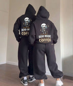 Two people standing indoors wearing matching black hoodies and sweatpants sets, shown from the back. Each outfit has a large white skeleton graphic and the text 'DEAD INSIDE COFFEE' on the back.