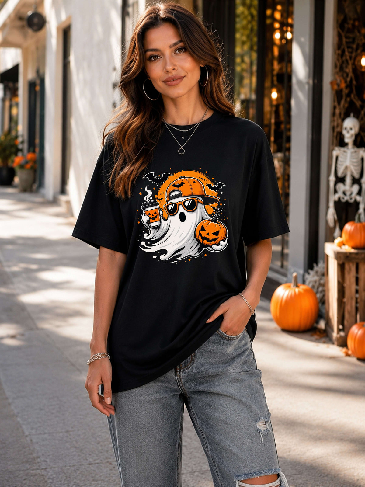 A woman wearing a black graphic t-shirt with a Halloween design featuring a ghost, jack-o'-lantern, and orange pumpkin, paired with distressed gray jeans, standing on a street decorated for the holiday.