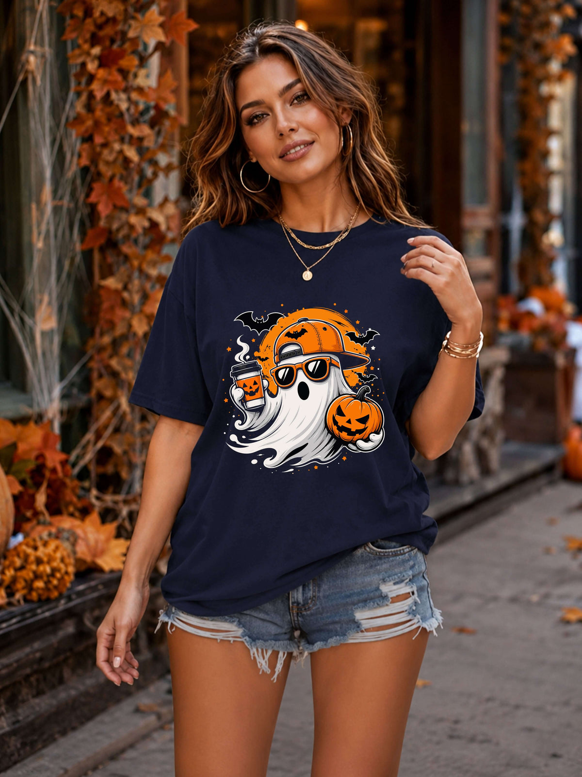A woman wearing a navy blue graphic t-shirt with a Halloween design featuring a ghost, jack-o'-lantern, and moon, paired with denim shorts, standing in an autumn-themed outdoor setting.