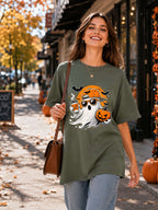 A woman walking on an autumn city sidewalk wearing an oversized olive green t-shirt with a Halloween graphic of a ghost, jack-o'-lantern, and bats. She is carrying a brown shoulder bag.