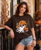 A woman wearing a dark brown oversized graphic t-shirt with a Halloween design, paired with light-wash denim shorts, posed outdoors in front of a fall-themed porch with pumpkins and autumn decorations.
