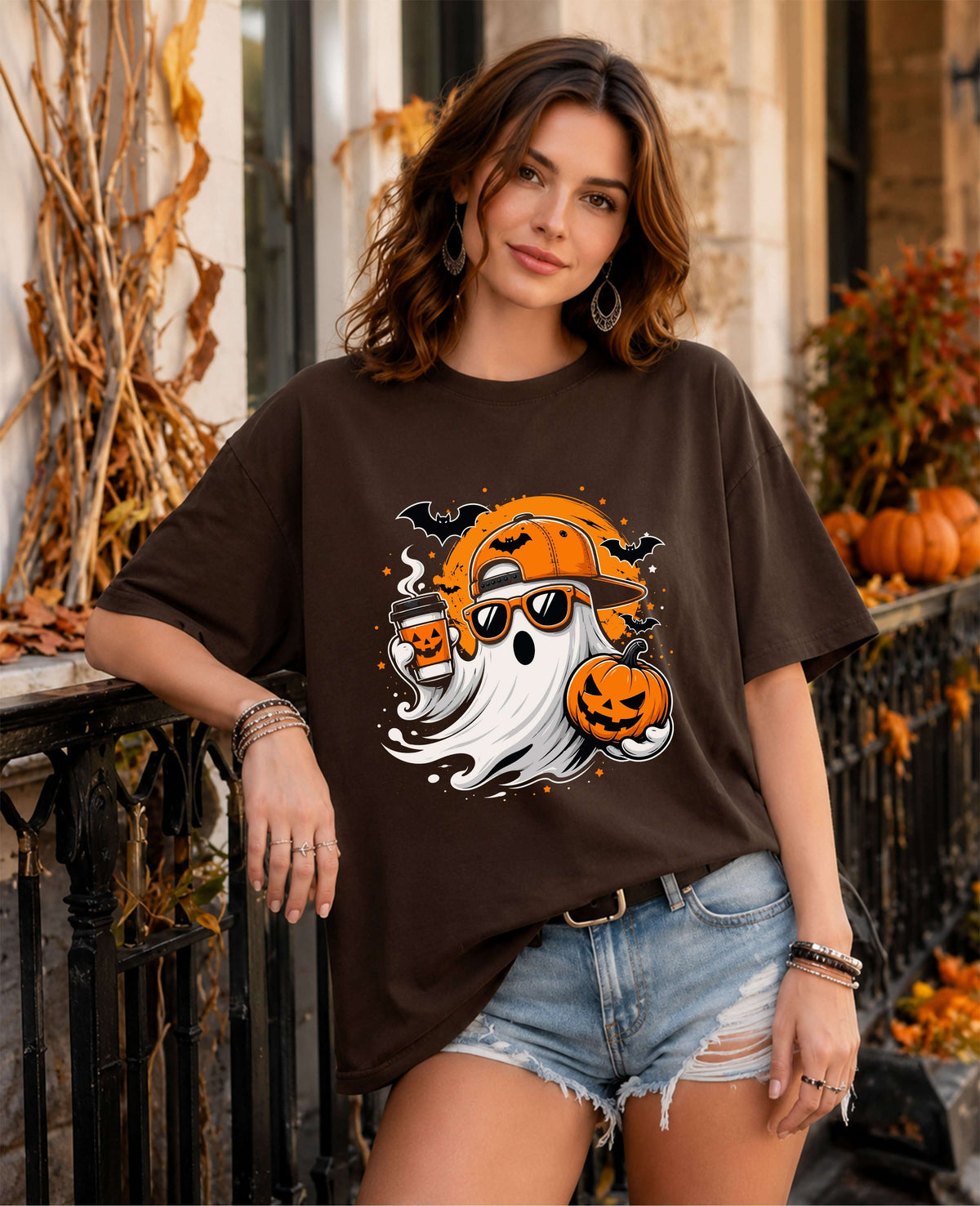 A woman wearing a dark brown oversized graphic t-shirt with a Halloween design, paired with light-wash denim shorts, posed outdoors in front of a fall-themed porch with pumpkins and autumn decorations.
