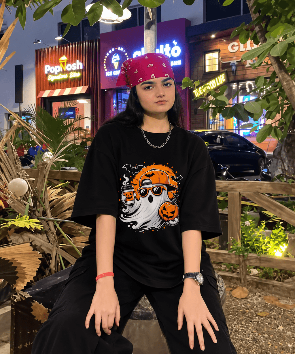 A person wearing a black oversized graphic T-shirt with a Halloween ghost and pumpkin design, black pants, and a red bandana, sitting outdoors at night in front of lit storefronts and foliage.