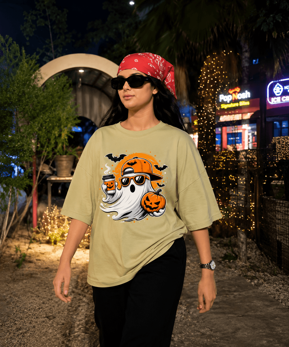 A woman wearing a beige graphic t-shirt with a Halloween print, a red bandana, and black pants, standing outdoors at night in front of a lit restaurant.